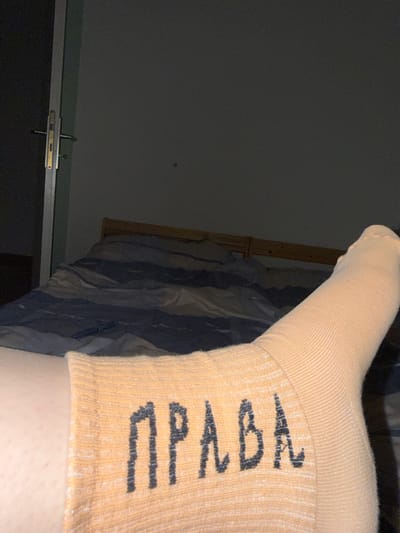 In an indoor setting, a person's leg, clad in a peach-colored sock with the Cyrillic letters "ПРАВА" printed in dark grey, is positioned in the foreground. The sock exhibits a ribbed texture. Behind the leg, a bed with blue and white striped bedding is visible. To the left, a doorframe with a silver handle and lock is present, set against a dark wall. The overall lighting suggests a dimly lit environment, possibly nighttime. This scene is not situated in Lufingen, Switzerland.