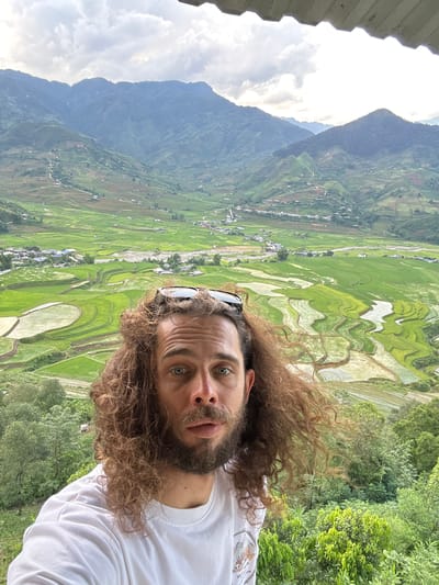 An outdoor selfie captured in Tú Lệ, Vietnam, features a man with long, curly brown hair, a beard, and light-colored eyes, looking directly at the camera with a slightly open mouth. He wears a white t-shirt and sunglasses pushed onto the top of his head. The man occupies the lower half of the frame, positioned against a panoramic view of a mountainous valley. The background showcases extensive terraced rice paddies, varying in color from vibrant green to reflective water, indicating different stages of cultivation. Numerous small structures and dwellings are scattered across the valley floor and along the lower mountain slopes, connected by visible dirt paths. Layers of densely vegetated mountains rise in the distance under an overcast sky with visible clouds. The immediate foreground below the man shows lush green foliage, while a section of a grey, corrugated roof is visible at the very top of the image.