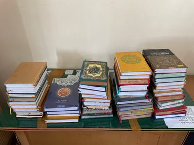 The image depicts a collection of books stacked on a wooden table with a green surface. The background is a light-colored wall with a faint crack running across it. In the foreground, on the left, is a tall stack of books with tan and white covers. Next to it, a dark blue book titled "Al Fattah Al Quran Khat Indonesia" is placed on top of a stack of smaller books. To the right of these, a stack of books with yellow and green covers is visible, with one featuring Arabic script. Further to the right, another stack of books with black and gold covers is prominent. The table surface has some light-colored tape and paper with text, part of which is visible. The scene is likely an indoor setting, possibly a library or study area in Tibang, Indonesia. There are no people or actions visible in the image.
