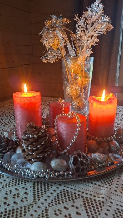 This image displays an indoor decorative arrangement situated on a table in Petriškė, Lithuania. A circular, silver-toned tray serves as a centerpiece, resting on a cream-colored lace tablecloth. On the tray are four red pillar candles; two are lit, one on the left and one on the right, exhibiting distinct orange-yellow flames. Surrounding the candles are multiple brown pinecones, strands of small silver beads, scattered gray pebbles, and translucent, irregularly shaped decorative crystals. A dark, spherical, openwork metallic ornament is also visible on the right side of the tray. Behind the tray, a clear glass vase holds several silver, glitter-coated spherical ornaments and artificial silver foliage, including a large openwork flower and a branched, frosted plant-like stem. The background consists of a wooden wall segment to the left and a dark fabric or curtain to the right. Illumination is provided by the burning candles. No individuals are present in the scene.
