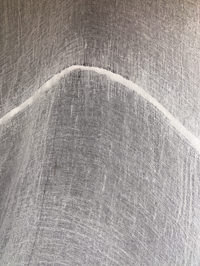 The image is a close-up, high-angle shot of a wrinkled, sheer white fabric. The fabric has a coarse weave, creating a textured, almost net-like appearance with many small holes. A raised, lighter white seam or band runs diagonally across the fabric from the upper left to the lower right, creating a wavy effect where it intersects the fabric's folds. The background is dark, suggesting the fabric is either hanging in front of a dark surface or the lighting is casting shadows. There are no people, animals, or specific landmarks visible in the image. The location in Райымбек, Kazakhstan cannot be determined from the visual information provided.