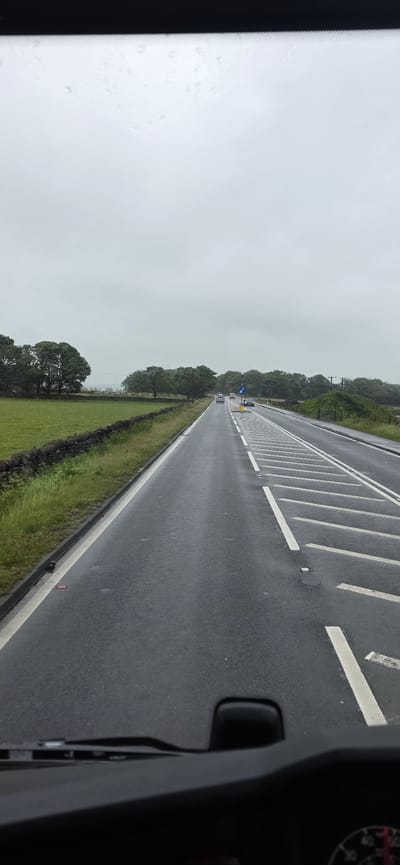 The image shows a grey, overcast day with light rain on a dual carriageway road, viewed from the perspective of a vehicle's dashboard. The road is wet and reflects the grey sky. There are white dashed lines on the road, indicating lanes, and some reflective markers. On the left side of the road, there is a low stone wall bordering a vibrant green grassy field and several large, mature trees. On the right side of the road, there is a verge with some vegetation and trees in the distance. Several cars are visible driving on the road. One car is in the left lane and two cars are in the right lane. A blue sign with a white arrow is visible on the right side of the road. The scene appears to be a rural or semi-rural setting in or near Barnsley, United Kingdom, likely a main road with some traffic.