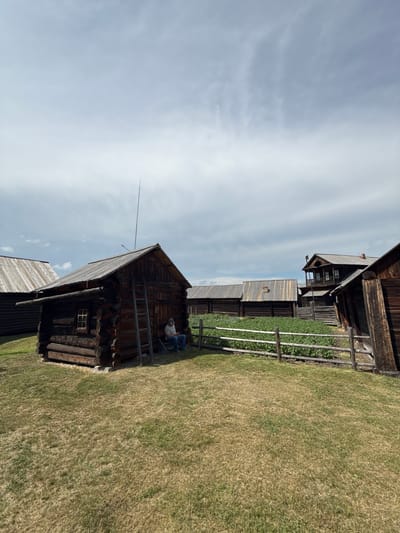 The image shows an outdoor scene in the city of Shushenskoye, Russia, likely at an open-air museum or historical settlement, given the presence of traditional wooden structures. In the foreground, dry, yellowed grass covers the ground. To the left, a rustic, dark wooden log cabin with a slanted roof and a small window is prominently featured. A wooden ladder leans against the side of this cabin. A person, wearing a light-colored shirt and blue jeans, sits on a wooden bench to the right of the cabin, appearing to be looking at a device in their hands.
Behind the cabin, a wooden fence with horizontal rails encloses a garden area filled with rows of green plants, possibly vegetables. Beyond the garden, several other wooden buildings of varying designs and sizes are visible, including a larger, multi-story structure with a balcony. The sky above is overcast with a mixture of gray and white clouds. The overall impression is a historical village or settlement setting under a cloudy sky.