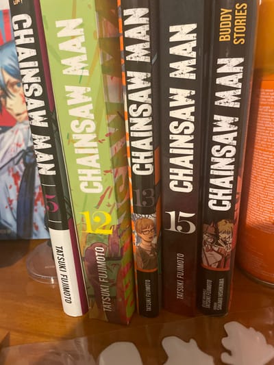 A stack of "Chainsaw Man" manga volumes is displayed. Volumes 12, 13, and 15 are distinctly visible. The spine of each manga showcases the title, author "Tatsuki Fujimoto," and volume number. The top volume is titled "Buddy Stories" and has writing that says "Sakaku Hishikawa". The bottom volume shows a fraction of the volume cover with a blue-haired character covered in blood.