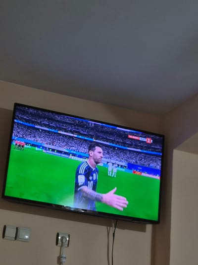 In the city of Kaleytsa, Bulgaria, a widescreen television is mounted on a light-colored wall. The television displays a soccer match, with a player in a blue and white striped jersey, identified as Lionel Messi by his appearance, in the foreground. He is on a bright green soccer field, with spectators visible in the background stadium seating, which is predominantly blue and white. A scoreboard in the upper right corner of the screen shows Cyrillic text and a score of 1-0. In front of the television, on the wall, are two light-colored light switches on the left and a white security camera with a black lens mounted on a short stalk to the right. The wall appears to be painted a neutral, light color. The overall scene depicts a living room or entertainment area where a soccer game is being watched.