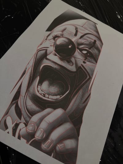 The image shows a close-up, overhead shot of a piece of paper with a black and white drawing of a screaming clown. The drawing features the clown's face, a red nose, wide-open mouth with teeth, and a hand holding something. Red lines are overlaid on the drawing, possibly indicating stencil lines for a tattoo. The paper is placed on a dark, shiny surface, possibly a table covered with plastic wrap, which has a textured, wet appearance. The scene is lit from above, casting subtle shadows. The image does not provide any information about the location in the city of Deventer, Netherlands.