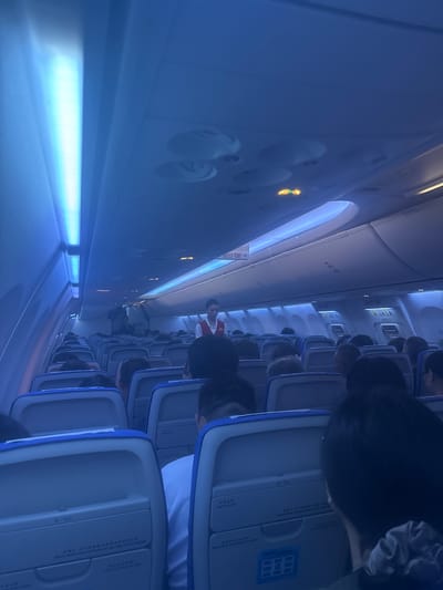 The image depicts the interior of an airplane cabin, likely during a flight, as indicated by the presence of rows of seats, overhead compartments, and cabin crew members. The lighting is predominantly blue, possibly due to ambient lighting or filters. Several passengers are seated, facing forward, with some partially visible. Two cabin crew members are visible; one standing near the front of the cabin, and another walking down the aisle. Signs, including "EXIT" signs in Chinese and English, are visible above the seats, suggesting this is an international flight or a flight with international signage standards. The overall scene is consistent with a typical airplane cabin environment, with no specific identifiable landmarks that would pinpoint its location within Xinwu District, China, or any other city. The image is taken from a seated passenger's perspective, looking towards the front of the cabin.
