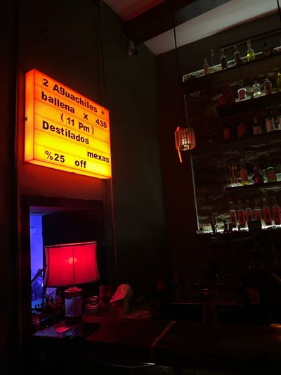 The image captures a scene inside a dimly lit establishment in Mazatlán, Mexico. A sign prominently displays the offer "2 Aguachiles + ballena x 430 (11 pm) Destilados mexas %25 off". The sign is illuminated with a bright yellow light. Behind the sign is an opening/window that leads to a darker space featuring a red lamp. The overall ambiance suggests a bar or restaurant, possibly after dark. Above, on the ceiling, are multiple shelves holding rows of bottles. The lighting is predominantly artificial and creates a stark contrast between the bright sign and the shadowy corners of the interior.