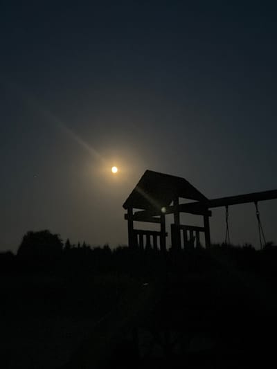 The image shows a silhouette of a swing set structure against a dark, hazy sky. The moon, appearing as a bright, luminous orb, is visible in the upper left quadrant of the image, casting a soft glow. A faint sunbeam effect emanates from the moon, creating a lens flare. The swing set structure, made of dark wood or metal, has a triangular roof and a series of horizontal bars, suggesting it is a play structure or perhaps part of a larger outdoor installation. To the right of the swing set, the dark silhouette of trees or foliage is visible, forming a dense, irregular line against the sky. The overall scene is dark and atmospheric, suggesting it is either dusk or night. The haziness in the sky could indicate fog, mist, or dust, contributing to the diffused light from the moon. There is no visible text in the image. The scene evokes a sense of quietude and perhaps a touch of mystery due to the low light conditions and the stark silhouettes. The location context of La Roche-en-Ardenne, Belgium, suggests an outdoor setting, possibly a park, garden, or rural area.