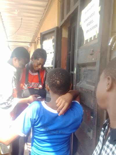 The image displays four individuals gathered near an entrance in Neni, Nigeria, under bright daylight. On the left, two males are observed interacting with a black portable electronic device. The leftmost male wears a white shirt with a dark pattern. The central male wears an orange "LAKERS" basketball jersey over a white undershirt and has braided hair; a black strap is visible across his chest. A third individual, viewed from the rear, wears a blue t-shirt with 'DR. SIR PATRICK AGU HOUSE' printed on the back, and rests their right arm on the central male's shoulder. A fourth individual, partially visible in profile on the far right, wears a black and white plaid shirt. The group is positioned in front of a building featuring a yellow-painted wall and a dark, worn double door. A white sign is taped to the right door, stating: "DMC ORIGINAL OFFICES", "WORKERS NEEDED URGENTLY.", and "(APPLY IN PERSON)". A corrugated metal roof overhang is visible above the scene on the left.