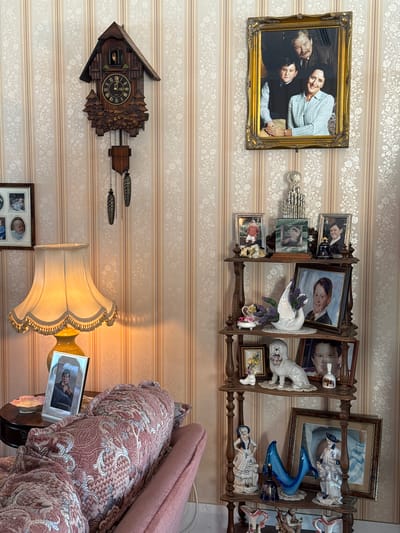 The image presents an interior scene with a pronounced focus on wall-mounted decorative items. The background is a wallpaper featuring stripes and floral patterns.

On the upper left, a gold-framed portrait of four people hangs on the wall. To the right and slightly higher, there is a wooden shelf filled with various framed photographs and figurines, including a white swan.

Below the shelf, a cuckoo clock with hanging weights and chains is prominently displayed. A lamp with a frilled shade illuminates the lower center of the image. Beneath the clock, another picture frame is placed on the wall.

To the lower right, part of a patterned armchair or sofa is visible, with a framed photograph resting on a small table next to it. The scene gives the impression of a domestic interior, likely a living room, densely decorated with personal items. The location, Nerima, Japan, is not explicitly confirmed by elements in the image.