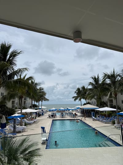 This is a photograph of a resort pool area, likely in Naples, Florida, United States of America, given the tropical vegetation. The image is taken from an elevated, covered vantage point, looking out over the pool and towards the ocean.
In the foreground, a large, rectangular swimming pool dominates the scene. The water is a vibrant turquoise. Several people are visible in and around the pool. One person is swimming in the deeper section of the pool, while others are in the shallower end, with one person appearing to be a child playing in the water. A pool lift mechanism is visible on the edge of the pool.
Surrounding the pool are paved deck areas with lounge chairs and umbrellas. Many lounge chairs are occupied by people relaxing. Buildings with balconies and windows line the sides of the resort area, and numerous palm trees are scattered throughout, indicating a warm climate. A white canopy structure with blue trim provides shade in the background, and it appears to be a bar or food service area.
In the distance, the ocean is visible under a cloudy sky. The overall atmosphere is one of a relaxed resort vacation. The sky is overcast with scattered clouds, suggesting the possibility of rain or a break in the sun. The lighting is diffused, typical of an overcast day.