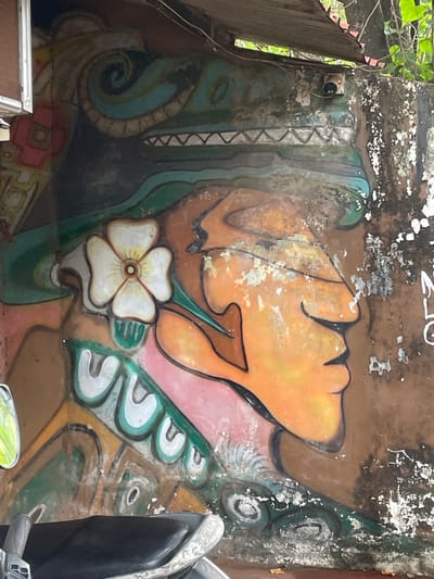 The image depicts a section of a weathered mural on a concrete wall in Vagator, India. The mural features a stylized profile of a face, predominantly in shades of orange and brown, with dark outlines. A large, white, four-petaled flower with a yellow center is positioned near the ear area of the face. Above the face, a green, serpentine design with jagged teeth suggests a mythical creature or headdress. Geometric patterns in red and yellow are visible on the left side of the mural, and a turquoise band with square-like elements runs across the top. The concrete wall exhibits significant wear and tear, with peeling paint and patches of exposed concrete, particularly on the right side. The scene is set outdoors, with foliage visible in the upper right corner. In the foreground, a portion of a black motorcycle seat and handlebar is visible, suggesting the mural is located in an area accessible by vehicles, possibly a roadside or parking area. A security camera is mounted on the wall above the mural. The overall impression is one of urban decay combined with vibrant, albeit faded, street art.
