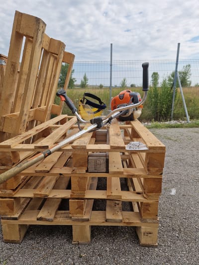 The image depicts an outdoor scene in Bahovitsa, Bulgaria, featuring several wooden pallets stacked on a gravel surface. A brush cutter with an orange engine and a yellow protective visor is positioned on top of the pallets. The brush cutter's shaft extends towards the left of the frame, resting on the wooden structure. In the background, a metal fence separates the area from a grassy field with scattered trees, under a cloudy sky. A small handwritten tag is attached to the side of the pallets.