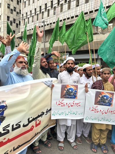 This media file captures a protest taking place in Karachi Division, Pakistan. A group of men, many wearing traditional Islamic attire and headwear, are seen holding green flags and banners. The banners display text in Urdu, with some reading "یہودی پرو پیگنڈا" (Jewish Propaganda) and "حب سنی علماء کونسل پاکستان، کراچی" (Love Sunni Scholars Council Pakistan, Karachi). Another banner features an image of a television logo with a chain and padlock over it.
The people in the image appear to be actively participating in the demonstration, with some raising their hands and others looking towards the camera. The setting suggests an outdoor urban environment, with a building facade visible in the background. The overall tone of the image is one of protest and dissent. The presence of green flags could indicate affiliation with a political or religious group. The specific text on the banners points to a protest against what is perceived as "Jewish propaganda" and possibly a call for support or attention towards the "Sunni Scholars Council." The time of day is not explicitly discernible, but the lighting suggests it might be daytime. The weather is not evident from the image.