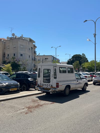 The image shows a street scene in Zvulun Regional Council, Israel. A white Toyota truck is parked along the side of the road, next to other parked cars. Buildings are visible in the background, with a clear blue sky above. Streetlights are also present. The road surface is asphalt, with some debris or fallen leaves visible near the curb.