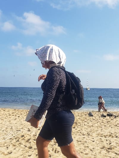 A woman in a patterned shirt, dark shorts, and a headscarf stands on a sandy beach. She has a black backpack and holds a small white book or album in her hand. Behind her is the ocean, and in the distance, a sailboat can be seen. Another person stands further down the beach. The sky is blue with scattered clouds.