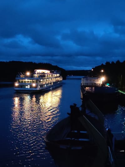 The image depicts a twilight scene on a body of water, likely a canal or river, near Devyatiny, Russia. Two vessels are visible. In the foreground to the left, a white passenger ship with multiple decks is illuminated by warm interior lights, casting reflections on the rippling water. The ship appears to be moving. To the right and further back, a dark cargo barge is also present, with some lights on its superstructure. The water is dark blue, reflecting the lights from the ships and the deep blue twilight sky, which is partially obscured by clouds. The shoreline is lined with dark trees, silhouetted against the fading light. The overall atmosphere is serene and tranquil.