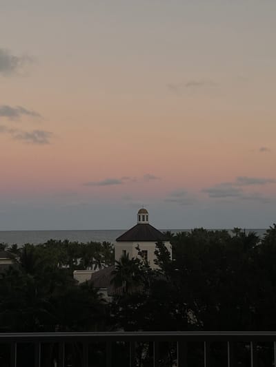 A picturesque scene showcases a building with a distinctive cupola, nestled amidst lush greenery. The structure is near a body of water, likely the ocean, which is visible as a horizontal line in the image. The sky above is a soft gradient of colors, possibly indicating sunrise or sunset. Part of a building with dark windows frames the right side of the photograph.