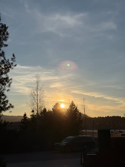 The image shows a sunset in Озёрное, Russia. Wispy clouds stretch across the sky, transitioning from a pale blue-gray to a golden hue as the sun nears the horizon. The sun itself is partially obscured by trees, creating a burst of light. The silhouettes of pine trees appear in the foreground. A dark-colored car and indistinct structures are visible on the right. In the distance, across a body of water, is a wooded shoreline.