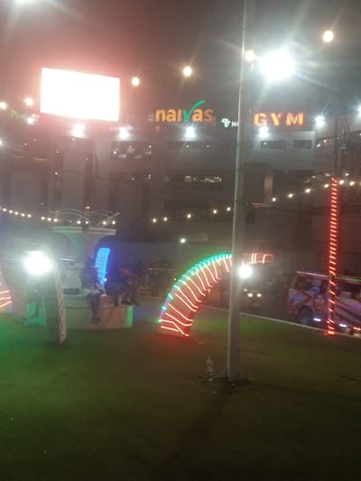 This image depicts a brightly illuminated night scene in Majengo ward, Kenya, focused on an outdoor commercial area. In the background, a multi-story building displays prominent illuminated signs: "Naivas" with orange lettering and a green checkmark, and "GYM" in red-orange characters. To the left of "Naivas," a large, brightly lit rectangular billboard is visible.

The foreground consists of a green turf area, presumably artificial grass. Within this area, several decorative light structures are present. To the left, a white illuminated arch leads to a blue arched light tunnel. Adjacent to this, a white stone bench or platform holds three seated individuals, their specific details obscured by distance and light. A central light pole extends vertically, and a clear plastic water bottle rests on the turf near its base. Further to the right, another large, arch-shaped structure is illuminated with alternating red and green lights.

On a paved surface to the right, two vehicles are visible. One vehicle, a van or SUV, features a distinct multi-colored custom paint job including a graphic of a face. Another vehicle with illuminated headlights is partially obscured behind the red and green light arch. Overhead, numerous string lights are suspended across the scene, contributing to the overall illumination.