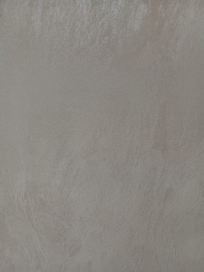 This is a close-up, textured image of a wall, likely an interior wall. The surface appears to be a matte finish with a subtle sheen, possibly stucco or a similar plaster. The color is a neutral, light beige or taupe. There are visible brushstrokes or trowel marks that create a slightly uneven, organic texture across the surface. Some areas appear slightly lighter or darker due to the play of light on the texture, creating subtle highlights and shadows. There are no people, objects, or discernable features that would indicate a specific location within the city of Hlynytsi, Ukraine. The image focuses solely on the wall's surface.