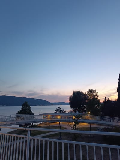 The image depicts an outdoor scene at dusk. The sky is a gradient of clear blue at the top, transitioning to soft pinks and oranges near the horizon, indicating sunset or twilight. In the foreground, a white metal railing runs horizontally across the bottom of the frame. Behind the railing, a pedestrian bridge curves across the lower half of the image, supported by visible pillars. The bridge is illuminated by warm yellow lights.
Beyond the bridge, a body of water, likely a lake, stretches to the horizon. On the opposite shore of the lake, a forested hill rises, dotted with lights from a settlement near its base. Several trees are visible in the mid-ground, some appearing windswept. The overall impression is a serene, natural landscape with man-made structures integrated into it. The location is identified as Sipplingen, Germany. There are no people visible in the image.