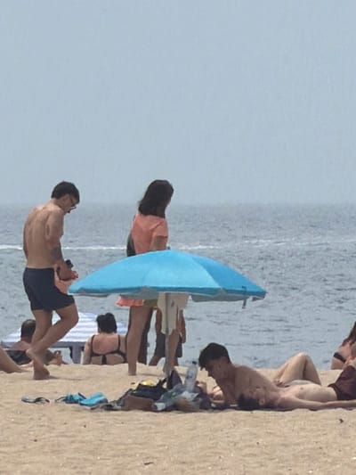 The image depicts a beach scene on a slightly overcast day, with the ocean in the background. In the foreground, a man in dark blue swim trunks stands facing left, his back to the viewer, looking downwards. To his right, a woman in an orange top and a light-colored skirt stands, facing away from the camera. A blue beach umbrella is prominent in the middle ground, partially obscuring a beach chair and another person lying down. Further back, a woman in a black bikini is seated on the sand. The beach itself is sandy, with various items of clothing and a water bottle scattered on it. The scene is likely the Praia da Baía in Espinho, Portugal, a popular urban beach known for its wide sandy expanse and proximity to the city.