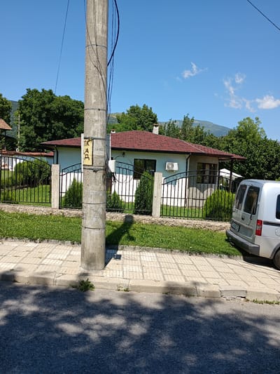 This is a sunny, daytime, exterior photograph taken in the city of Zlatitsa, Bulgaria. The scene depicts a residential street with a single-story house, a fence, a utility pole, and a silver van parked on the side of the road.

The house has a light blue facade, a red tiled roof, and several windows. An air conditioning unit is visible mounted on the exterior wall. A black metal fence encloses the property, with manicured green bushes and small evergreen trees planted behind it.

A concrete utility pole stands prominently in the foreground, with various wires and cables attached. A yellow sign with Cyrillic lettering is affixed to the pole. The street in front of the property is paved with asphalt and features a concrete sidewalk with a distinct pattern of square tiles. The curb appears to be damaged in places.

A silver van, possibly a passenger van or a small delivery vehicle, is parked on the right side of the image, facing away from the viewer. The rear of the van and part of its license plate are visible.

The background features lush green trees and a distant, hazy mountain range under a clear blue sky with a few scattered white clouds. The overall impression is a quiet, suburban setting on a warm day. No people are visible in the photograph.