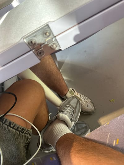 The image shows a close-up view of two pairs of legs and feet, likely inside a train compartment. One person's legs are visible wearing denim shorts and grey athletic shoes with white socks. A white charging cable is looped around their left leg. The other person's legs are partially visible, wearing white athletic shoes with orange accents and light-colored pants or shorts. The legs are positioned near each other, suggesting they are sitting in adjacent seats. The background includes a light-colored, textured surface above, possibly a table or tray, and a grey floor. The overall scene suggests a travel setting. There is no information in the image to indicate the location is Narbonne, France.