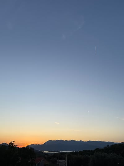 The image captures a sunset scene over Pelinovo, Montenegro. The sky displays a gradient of colors, transitioning from a soft orange near the horizon to a clear blue at the top. Two faint contrails from aircraft are visible in the sky. In the foreground, silhouetted trees and the rooftops of a few houses are visible. Beyond them, a range of mountains forms the backdrop, with a body of water nestled between the mountains and the treeline. The overall atmosphere is serene, depicting the end of the day in a coastal mountainous region.
