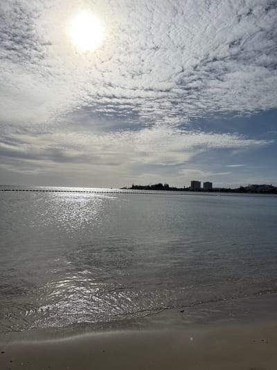 The image captures a coastal scene in Noumea, France, specifically a bay resembling Anse Vata or Baie des Citrons. The foreground consists of a strip of light-colored sand at the water's edge, exhibiting a wet line from the gently receding waves. The middle ground is occupied by calm, clear water, reflecting a strong, overexposed sun positioned in the upper left of the frame, creating a bright, shimmering glare across the water's surface. A dark line of buoys, connected by a rope or chain, extends horizontally across the water in the mid-distance, marking a boundary. The distant horizon reveals a landmass with a mix of dark green vegetation and multiple urban structures, including two prominent, multi-story buildings, likely hotels or apartments, visible towards the right. The sky is largely covered by a dense layer of altocumulus or stratocumulus clouds, with some patches of lighter blue visible, particularly towards the right. No individuals, animals, or active subjects are present in the scene.