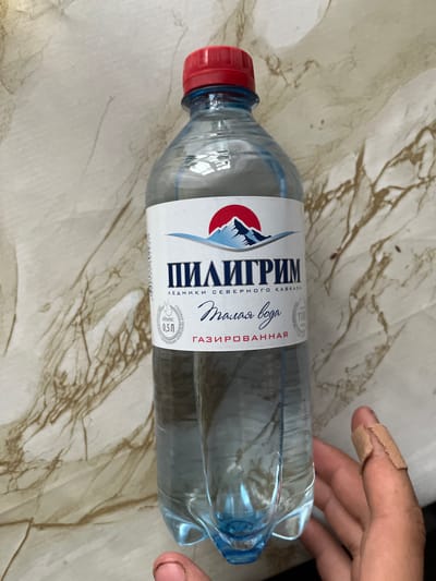 The image shows a clear plastic bottle of water with a red cap, lying on a patterned tablecloth. The label on the bottle is predominantly white and blue, featuring a mountain landscape with red accents. Visible text on the label includes "ПИЛИГРИМ" in large, bold letters, and below it, "СЕВЕРНОГО КАВКАЗА". To the right of this, in a cursive font, is "Талая вода" and "ГАЗИРОВАННАЯ". A circular emblem with text like "СТОЛОВАЯ ПРИРОДНАЯ" and "0,5Л" is also present.
A person's hand, with a bandage on the index finger, is visible on the right side of the image, near the top of the bottle. The hand appears to be gently touching or holding the bottle. The scene is indoors, as suggested by the tablecloth and lighting. No specific time of day or weather conditions are discernible. The overall impression is casual, possibly someone about to drink or having just picked up the bottle. The setting appears to be domestic, given the tablecloth. The location context suggests the water might be sourced from the Minyar region of Russia.