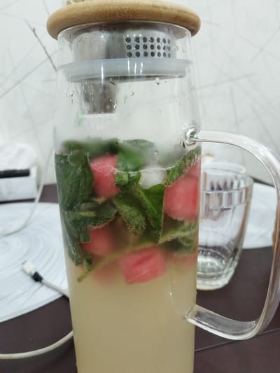 A close-up shot shows a clear glass pitcher filled with a light-colored beverage. The drink is infused with cubes of red fruit, likely watermelon, and fresh green mint leaves, creating a vibrant and refreshing appearance. Ice cubes are also visible, contributing to the cold beverage. The pitcher has a wide glass handle and a distinctive bamboo lid with a silver infuser. Condensation droplets are visible on the outside of the glass, suggesting the drink is cold.

Behind the pitcher, a clear glass with ridged detailing is partially visible, hinting at a second serving or that the pitcher is prepared for use. The background consists of a patterned surface, possibly a countertop or table, with a white and gray tiled wall in the distance. A white cable with a black connector is lying on the patterned surface to the left of the pitcher.

The lighting suggests it might be daytime, possibly indoors given the indoor setting cues like the tiled wall and furniture. There are no people visible in the frame, and no text is present in the image. The overall impression is one of a cool, homemade beverage ready to be served, likely in a home kitchen or dining area. The composition focuses on the refreshing ingredients within the pitcher.