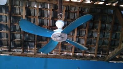 A blue and white ceiling fan with four blades is suspended from a wooden ceiling structure. The fan is centrally located and appears to be stationary. The ceiling itself is constructed of wooden beams and what appear to be corrugated roofing panels. The overall environment suggests an outdoor or semi-outdoor covered space. The lower portion of the image shows a bright blue wall. The location is identified as Banyusri, Indonesia.