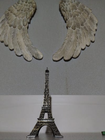 The image presents a static composition of decorative objects, viewed from a slightly elevated perspective. Two ornate, wing-shaped figures, resembling angel wings, dominate the left side of the frame. They are an off-white or light beige color with visible texture detailing individual feathers, and appear to be crafted from a solid material like plaster or resin. One wing is positioned in the upper left, and the other in the lower left, creating a symmetrical arrangement. Both wings cast distinct shadows on the underlying surface.

To the right of the wings, a miniature metallic replica of the Eiffel Tower stands vertically. The figurine is dark grey or bronze, featuring intricate structural details of the famous landmark's latticework. It is positioned upright on a lighter, plain off-white surface that contrasts with the textured grey surface beneath the wings. A faint seam or corner is visible where these two surfaces meet on the right side of the image.

The background comprises two distinct areas: a textured, light grey surface on the left, and a smoother, lighter grey or white surface on the right. The lighting suggests an indoor setting, with soft shadows indicating a light source from above or the side. There is no activity or event taking place, as the objects are static decorations. No people are visible, and there is no discernible text on any of the objects or the background. The scene is situated in Roza, Bulgaria.