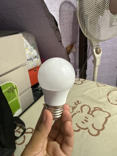 A close-up shot shows a hand holding a white LED light bulb. The background features a fan and a wall with a textured pattern. The scene appears to be indoors, possibly in a residential setting. The lighting is dim, with the bulb being the primary light source. No specific location in Manila, Philippines is identifiable from the image.