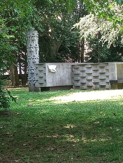 In the city of Olot, Spain, a concrete monument stands in a grassy park. The monument features a tall, cylindrical stone pillar decorated with relief carvings of small figures, possibly children, arranged vertically. Adjacent to the pillar is a rectangular concrete structure with a textured surface composed of repeating geometric patterns. A white, abstract relief of a bird's head is affixed to this structure. The monument is surrounded by lush green grass, with scattered leaves and dappled sunlight indicating an outdoor setting under trees. The background is filled with dense green foliage.