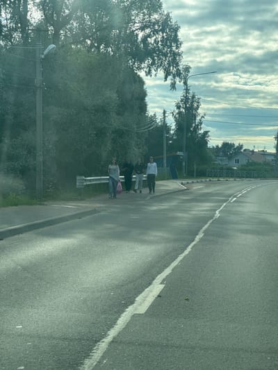 The image shows a street scene in Annino, Russia. The foreground features a paved road with a white line marking. The background is dominated by lush green trees and a cloudy sky. Several people are visible standing on the side of the road, some appearing to be waiting. One person is wearing a white shirt and dark pants, while others are dressed in darker clothing. One individual is carrying a pink bag. There are streetlights and power lines visible, suggesting an urban or suburban environment. The sunlight filtering through the clouds indicates it is daytime, likely late afternoon or early evening given the soft lighting and shadows. The overall atmosphere appears to be calm and ordinary, depicting everyday life on a road in a residential area. There are some buildings visible in the distance, nestled among the trees. No text is clearly legible in the image.