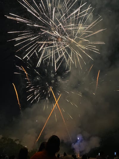 The image captures a nighttime fireworks display over Canet de Mar, Spain. The sky is filled with numerous streaks of light from exploding fireworks, primarily in white and gold hues, radiating outwards from various points. Some fireworks create a starburst effect, while others appear as long, trailing lines. Wisps of smoke from the explosions drift through the dark sky, obscuring some of the details. In the foreground, silhouetted against the bright pyrotechnics, are the heads and shoulders of a crowd of people watching the display. Trees are visible on the far left, and the lower portion of the image shows a gathering of spectators, some holding up phones to capture the event. The overall atmosphere is festive and celebratory, typical of a public fireworks show.