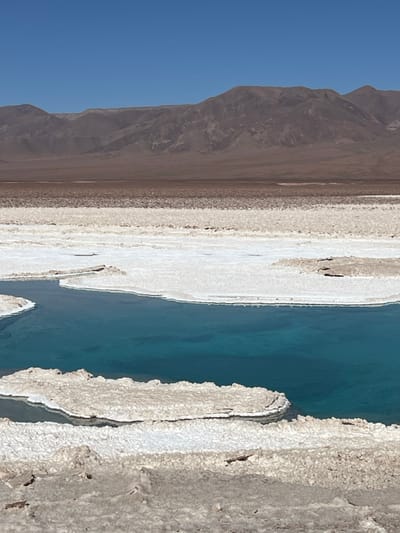 The image shows a landscape with a stark contrast between terrain types. On the left, mountains rise against a clear blue sky. In the center, a light-colored, potentially saline or mineral-rich area stretches alongside a body of water with a vivid turquoise hue. The right side features a rough, textured ground, possibly containing salt formations. The scene conveys an arid environment, where geological processes have shaped the landscape over time.