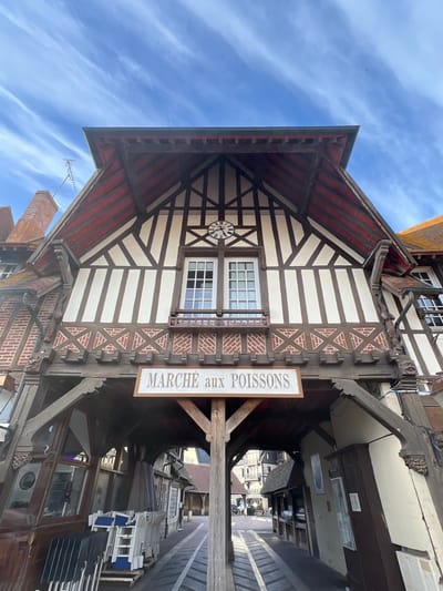 The image depicts the Marché aux Poissons (Fish Market) in Deauville, France, under a bright blue sky with scattered white clouds. The building features traditional Norman architecture with half-timbered elements, white stucco walls, and a dark red roof. A sign above the entrance reads "MARCHÉ aux POISSONS." The market appears to be an open-air walkway with stalls lining both sides. Several chairs and tables are stacked on carts, suggesting the market is either closed or preparing to open. The perspective is low-angle, emphasizing the scale of the building.