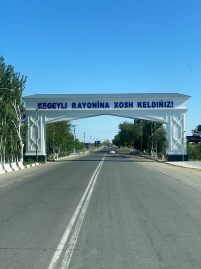 The image captures a wide-angle, eye-level view of an arched gateway spanning a paved road leading into the city of Khalkabad, Uzbekistan. The gateway, constructed of white material with decorative columns, bears blue lettering in Cyrillic script that reads "KEGEYLI RAYONINA XOSH KELDINIZ!". The road is asphalt, marked with double white lines in the center and a white striped barrier along the left side. Trees line both sides of the road, with a clear blue sky overhead. A few cars are visible on the road, suggesting ongoing traffic. The overall scene depicts a welcoming entrance to a district within the city.