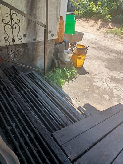 An outdoor scene in Talgar, Kazakhstan shows a rough, unpaved area with a few objects and structures. In the foreground, a dark metal grate is angled downwards, suggesting it might be a drainage grate or part of a walkway. To the right of the grate are several rough, dark wooden planks, also angled.
Behind the grate and planks, a weathered concrete wall is visible, with a white tarp-like material draped over it. A decorative black metal railing with a scrollwork design is attached to the wall.
Several containers are scattered around the area. To the left of the railing, there is a large, translucent orange plastic barrel. Next to it, a bright green plastic barrel is situated inside a weathered grey wheelbarrow. Below these barrels, on a concrete block, sits a small white bucket with a lid. To the right of the white bucket is a large, translucent yellow plastic container, possibly a jug or milk can, with a metal handle. Some green grass grows around the base of these containers and the concrete blocks.
The ground is a mixture of dirt and concrete, with tire tracks visible in the distance. Lush green foliage and trees are present in the background, suggesting a somewhat rural or semi-urban setting. The lighting indicates it is daytime, with shadows cast by the objects.