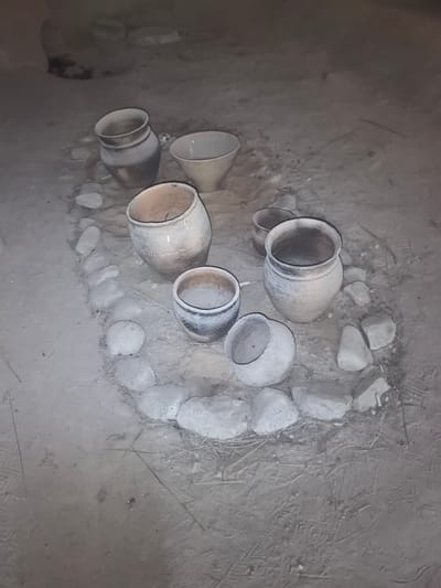 In Камянюцкі сельскі Савет, Belarus, a collection of ancient, earthenware pots and bowls are arranged in a circular pattern on a dirt floor. The pottery varies in size and design, with some exhibiting intricate etchings and decorative patterns. The pots appear to be aged, showing signs of wear and tear, with a rough, textured surface. The objects are placed on a floor made of compacted dirt, with several stones scattered around the pottery, possibly forming a border or support structure. The overall impression is that of an archaeological site or a display of historical artifacts, perhaps in a museum or an outdoor exhibit. The lighting is dim, casting subtle shadows and highlighting the textures of the pottery and the surrounding ground.