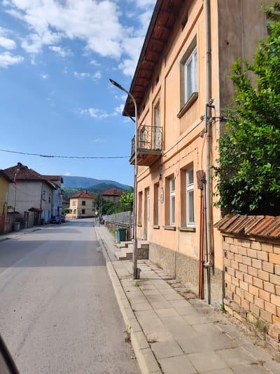 This is a street view in Lesidren, Bulgaria, on a sunny day with a bright blue sky and scattered white clouds. The perspective is from a vehicle looking down a paved street lined with buildings. On the right is a two-story building with an orange facade, featuring multiple windows, a small balcony with an ornate railing, and a tiled roof. A green tree grows next to this building, and a brick wall with a tiled top separates it from the sidewalk.
The street is empty of people but has parked cars visible in the distance. Buildings on the left side of the street are also two-story structures with tiled roofs. One building on the far left flies a Bulgarian flag. The street has a sidewalk on both sides, with some trash bins and a lamppost visible. In the background, rolling green hills form the horizon. The scene depicts a quiet, possibly rural, Bulgarian town.