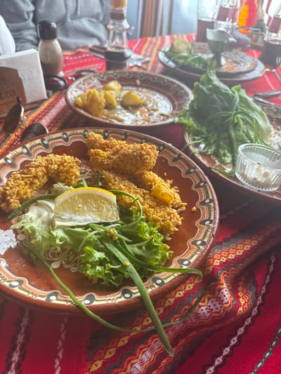 The image depicts a dining table within a restaurant located in Mala tsarkva, Bulgaria. The table is covered with a vibrant red tablecloth featuring traditional Bulgarian folk patterns in white, yellow, orange, and green.

In the immediate foreground, a brown ceramic plate with a distinctive patterned rim holds several pieces of golden-brown, breaded food, garnished with a yellow lemon wedge, green lettuce, and thin green shoots, likely spring onions. Adjacent to this, another similar ceramic plate contains remnants of consumed food, specifically yellow, possibly roasted or fried potatoes. To the right, a third identical plate holds a portion of fresh green lettuce and a small, clear glass ramekin filled with a white sauce or dressing.

In the blurred background, a person wearing a grey top is partially visible. Other objects on the table include a brown condiment shaker, a taller clear bottle containing a dark liquid, silverware, and blurred drinking glasses or bottles, one of which appears to contain a red beverage. A pair of sunglasses rests near a white napkin holder on the left side of the table. The scene presents a partially consumed meal in a restaurant setting.