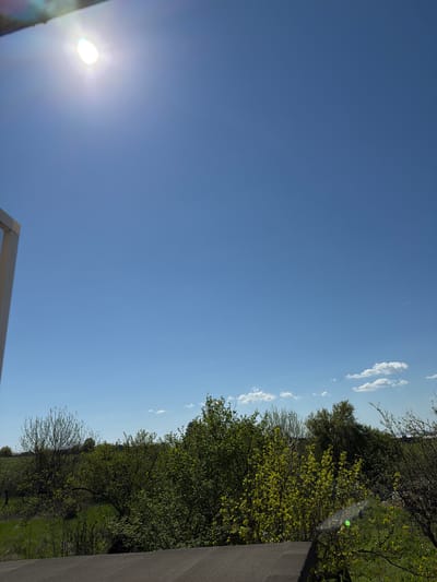 The image depicts a daytime scene with a clear blue sky, a bright sun, and trees. Thin clouds are visible near the horizon. The location is potentially Rosen, Bulgaria as requested, but that is not verifiable from the image alone. The scene includes elements from nature with trees lining the right side and the sun. A portion of a building structure is visible in the bottom left and bottom center of the frame.
