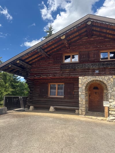The image shows a rustic, wooden chalet-style building with dark brown logs. The roof is made of wood planks with exposed beams and is partially visible against a bright blue sky with scattered white clouds. The building has multiple windows, some with closed wooden shutters. An arched stone entryway leads to a wooden door. A blue rectangular sign with the number "98" is affixed to the wall above the door. To the left of the building, there is a dark wooden railing and trees. The ground in front of the building is a gravel pathway. A mailbox is positioned to the right of the arched doorway. The overall impression is of a rural mountain dwelling. This building is located in the municipality of Les Rousses, France.