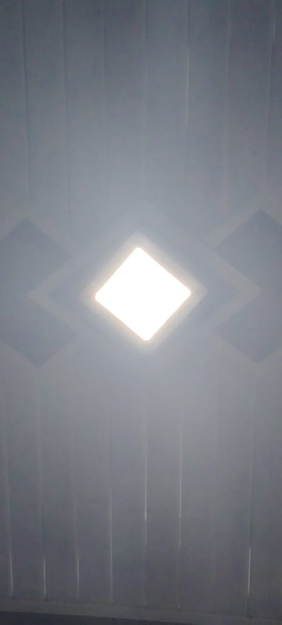 A downward-facing, close-up shot of a recessed, square-shaped ceiling light fixture in Jijiem, Indonesia. The light is emitting a bright, white light. The ceiling is paneled with vertical grooves and features concentric square recesses around the light fixture.